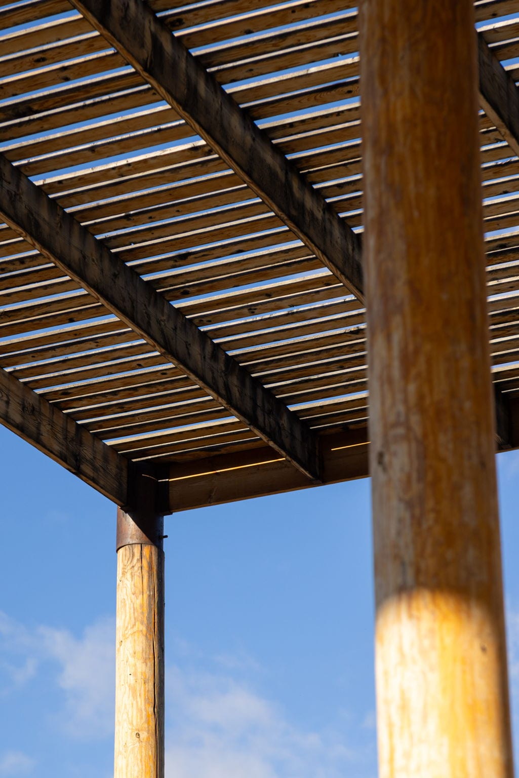 The wood lattice canopy of the Ramada House from below, with two round timber posts in the foreground and the horizontal beam-and-slat structure receding into perspective against blue sky.