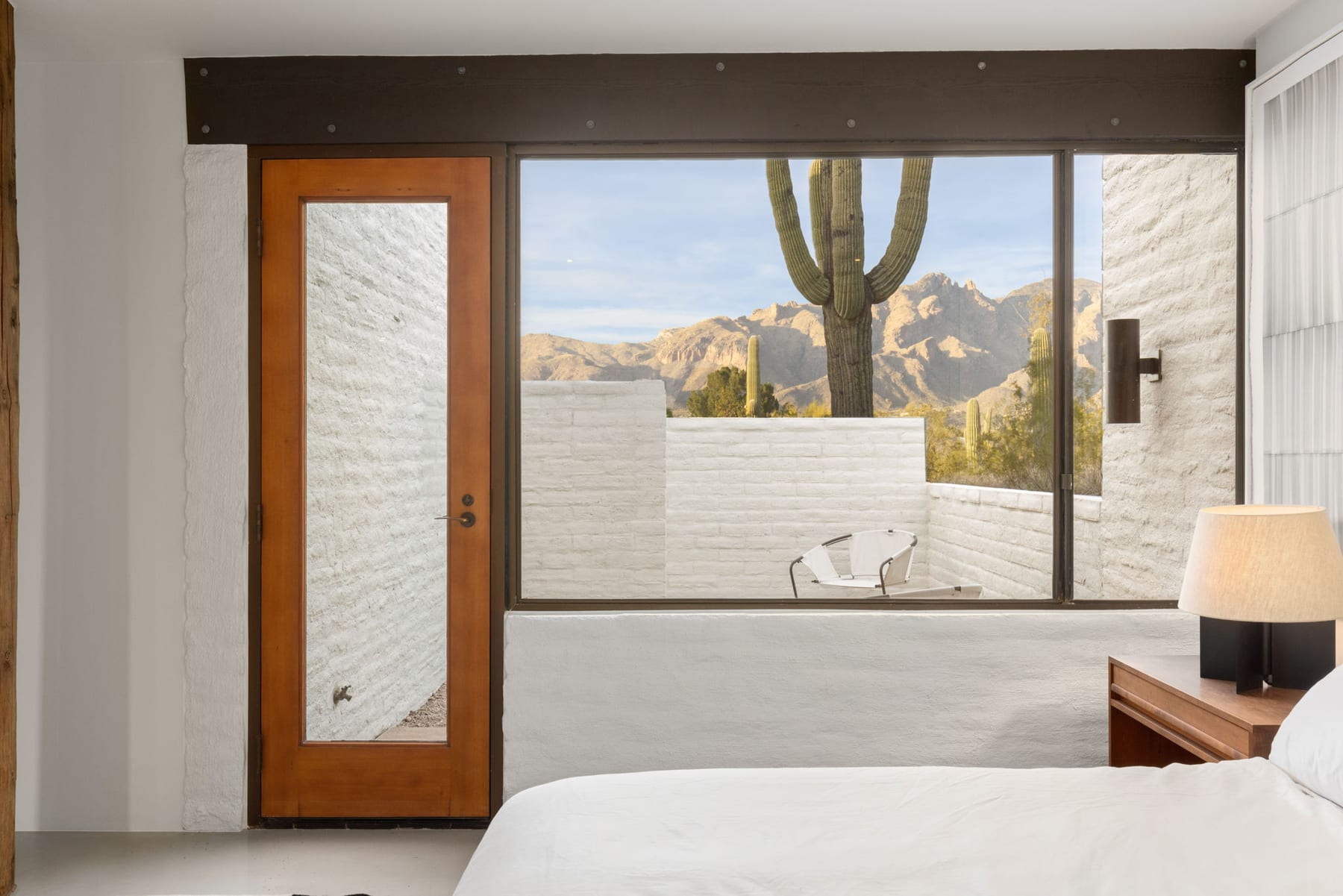 A bedroom interior at the Ramada House — a wood-framed door with mirrored panel at left, and a window at right looking into a small private courtyard with a saguaro and the Catalina Mountains beyond.
