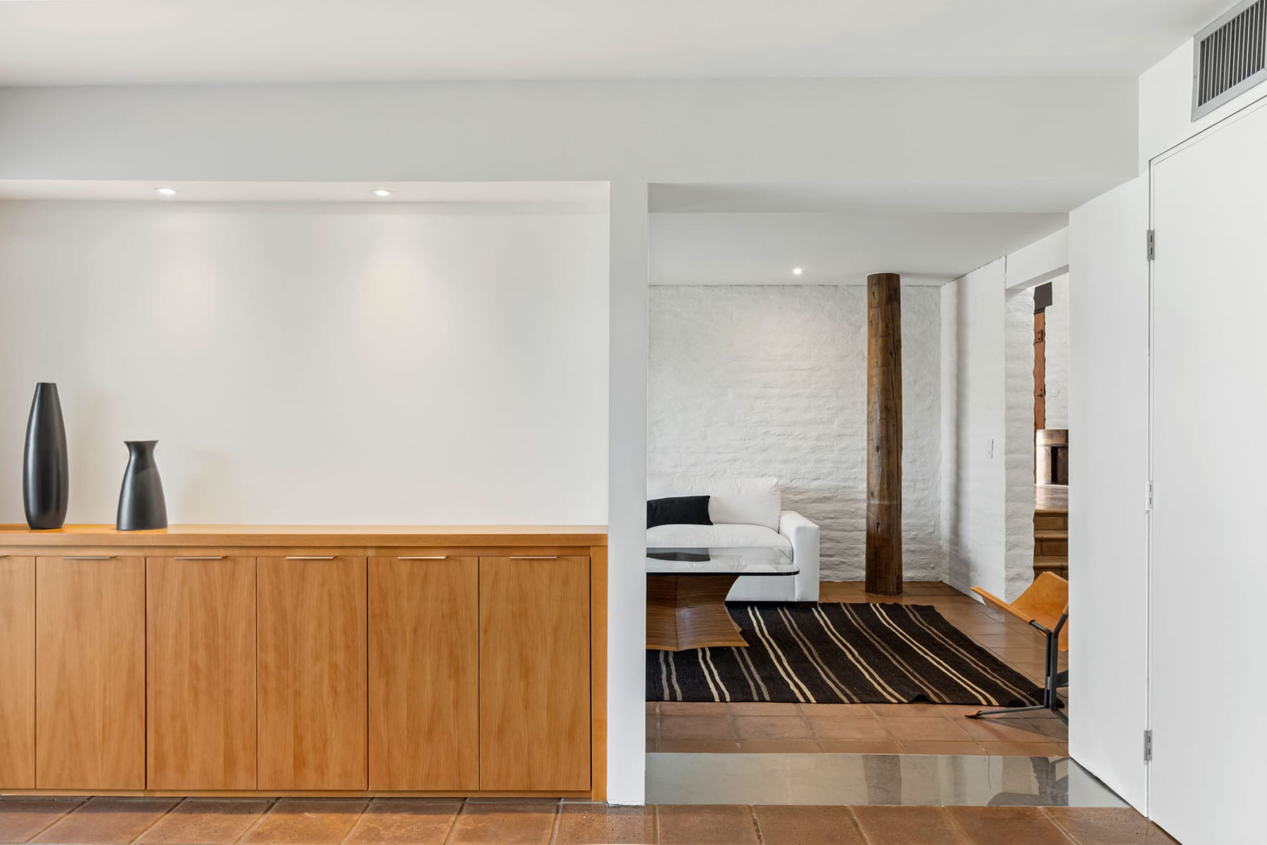 An interior view at the Ramada House — wooden built-in cabinetry with two ceramic vases in the left foreground, a doorway at center opening into a sitting area beyond with white sofa, striped rug, and one of the ramada&rsquo;s round wood posts piercing through to the floor.
