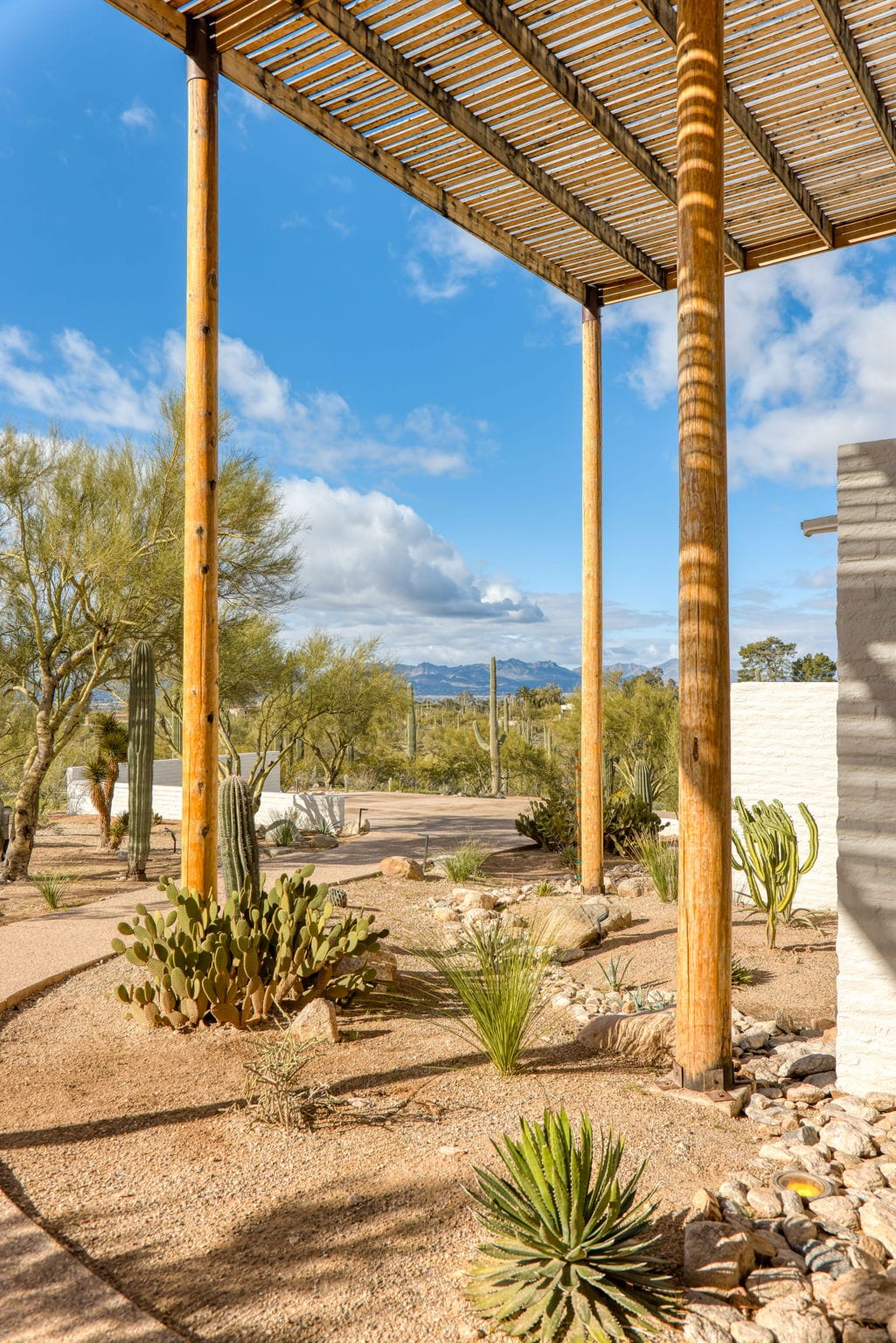 A view from under the Ramada House&rsquo;s lattice canopy — three round wood posts at vertical thirds, a gravel and cactus garden in the middle ground, and distant desert and mountain views beneath a sky of scattered clouds.