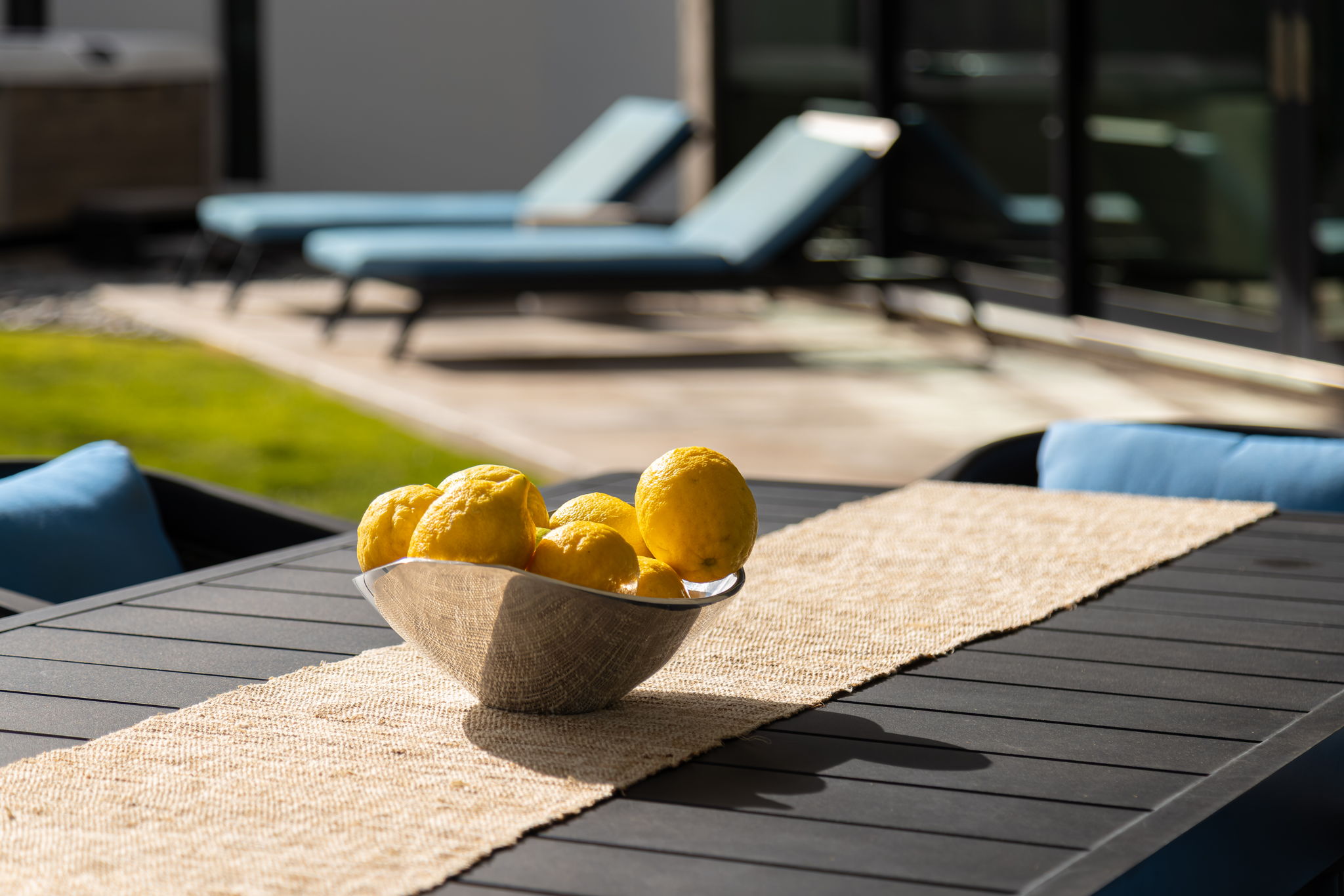 A bowl of sunlit lemons on a dark outdoor table, with the pool deck and chaise lounges defocused in the background.