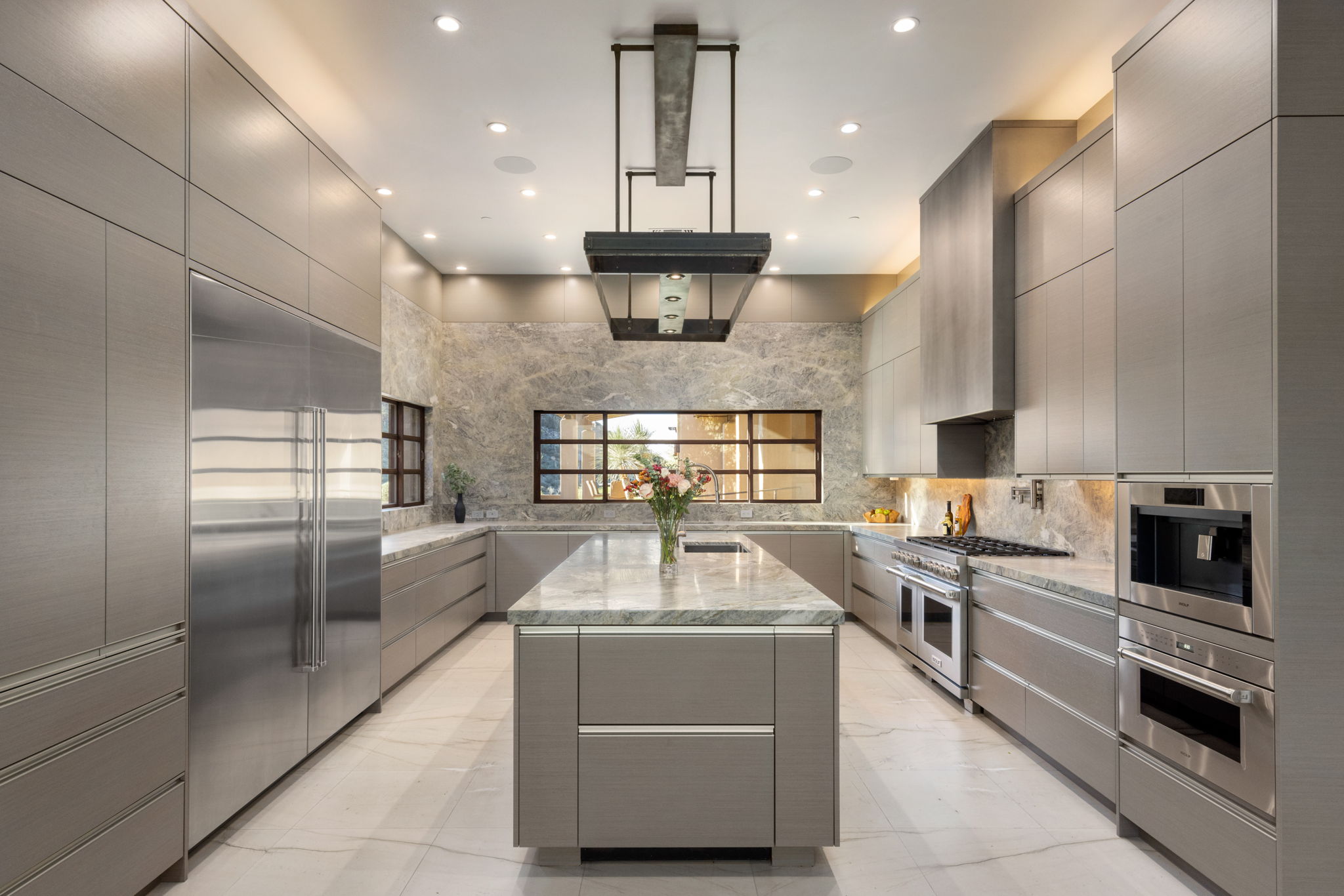 Kitchen interior &mdash; European-profile cabinetry in a cool brushed gray finish flanking both walls, a full-height granite slab backsplash, a sculptural steel and concrete range hood suspended from above, professional stainless appliances, marble tile floor.