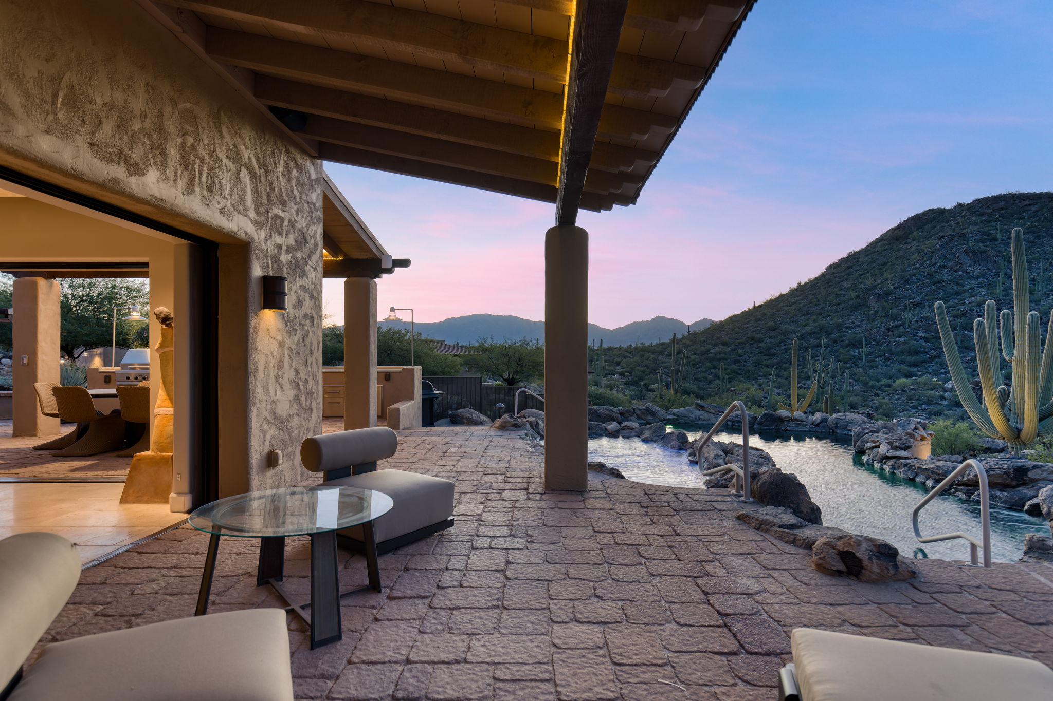 Covered outdoor living area at blue hour &mdash; a concrete column in the foreground, the pool threading between granite boulders into the desert beyond, a saguaro-covered hillside rising against a pink and blue dusk sky, warm interior light visible at left.