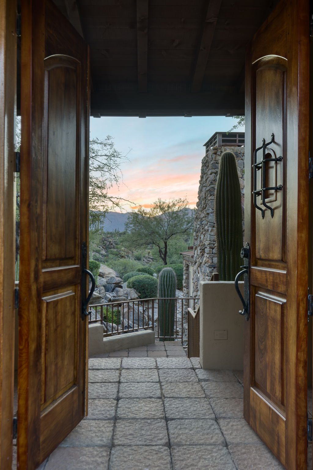 The designed arrival sequence at the compound &mdash; heavy walnut doors with wrought iron hardware open to stone steps descending toward saguaros, boulders, a fieldstone tower, and a pink and blue sunset sky.
