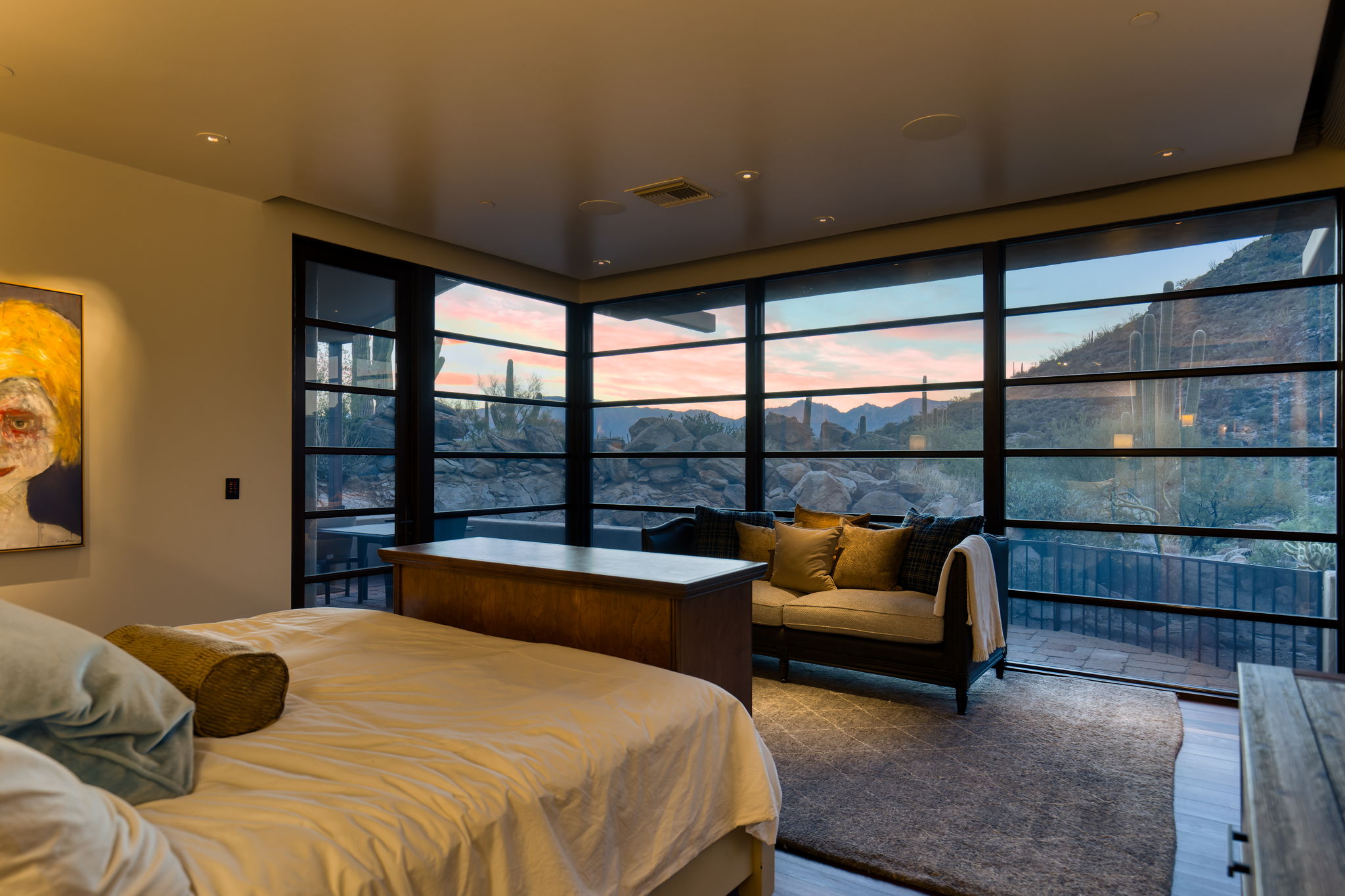 Primary bedroom at dusk &mdash; two full walls of steel-framed glass at the corner of the room reveal a saguaro-studded hillside with granite boulders and a pink and blue sunset sky, a chaise longue positioned toward the view.
