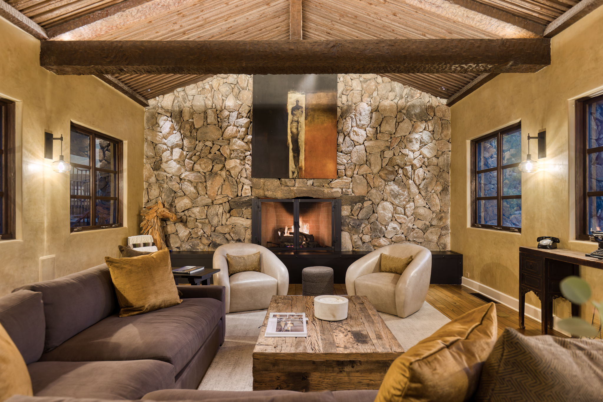 Living room interior &mdash; a full-height fieldstone fireplace wall with open fire, flanked by arched window openings with views to desert boulders, vaulted ceiling with rough-sawn wood planking and heavy timber ridge beam, warm amber lighting.