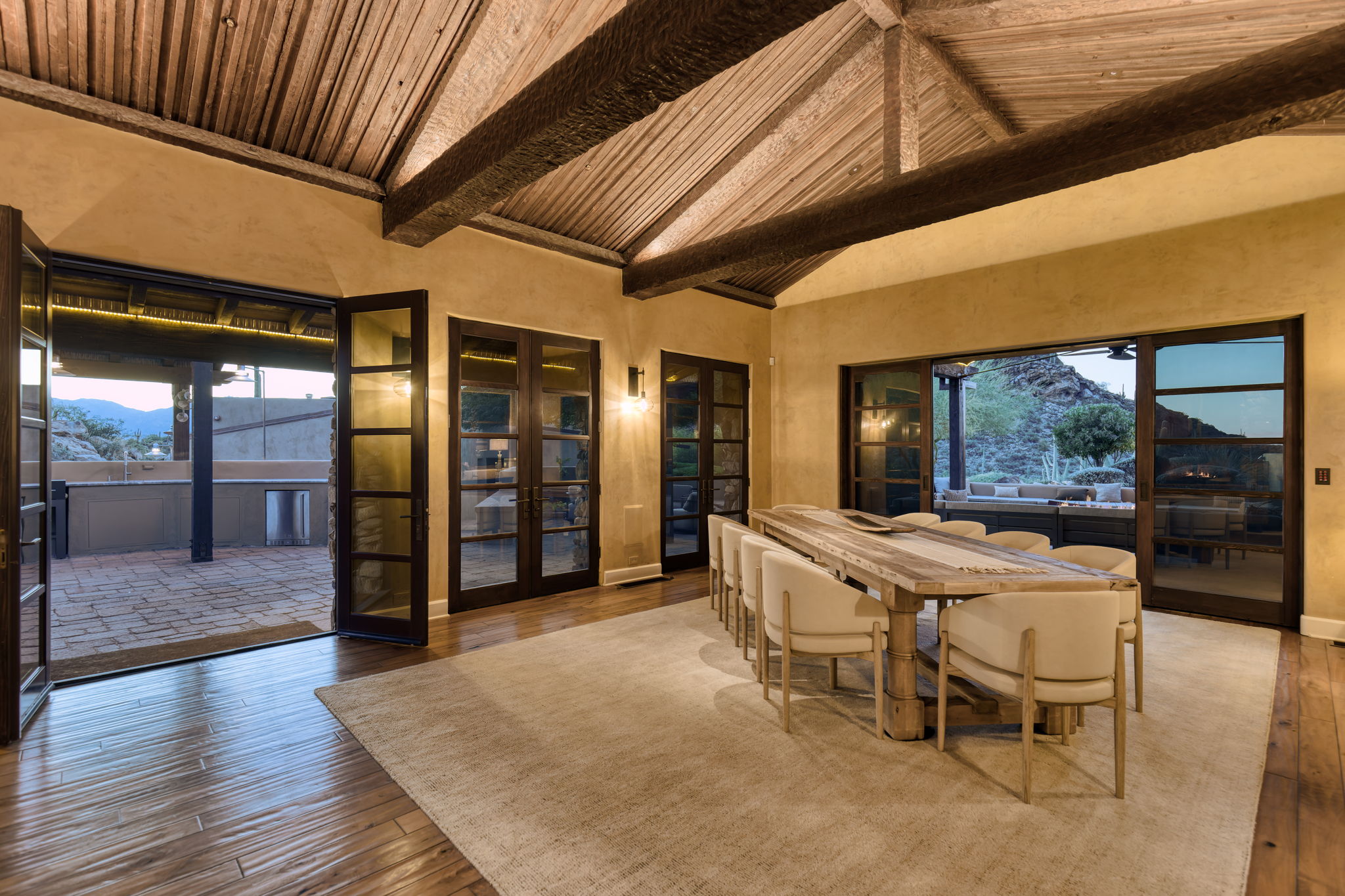 Dining room at blue hour &mdash; vaulted ceiling with rough-sawn wood planking and heavy timber beams above a long harvest table and upholstered chairs, french doors open to the terrace and desert beyond on two sides, warm lamp light against a deepening dusk sky.