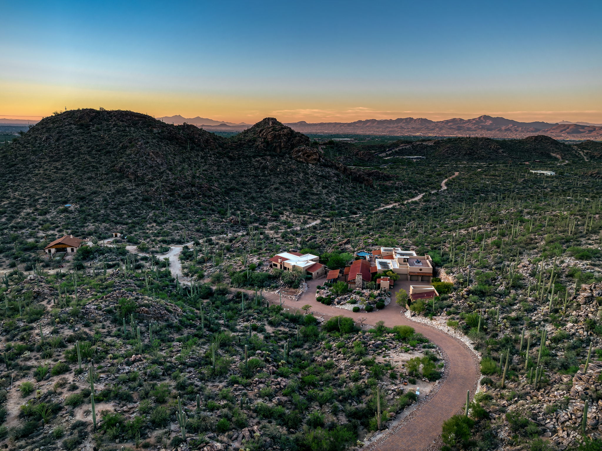 Aerial view at dusk of the Saguaro Ranch Retreat — a multi-structure compound set small in a vast Sonoran desert landscape, surrounded by saguaro-covered hills, with the Catalina Mountains on the horizon.