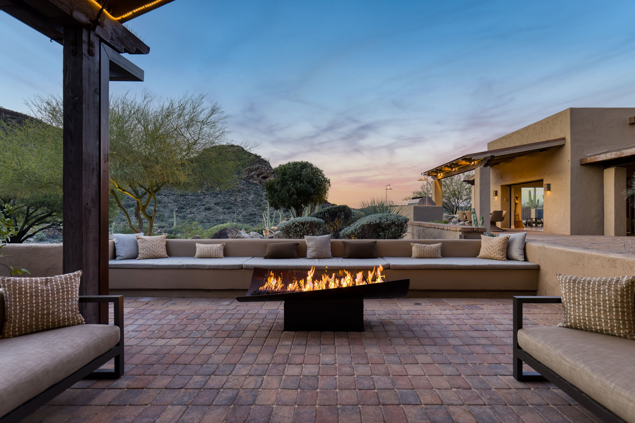 Fire terrace at dusk &mdash; a long built-in seating bench curves around a low rectangular fire feature with open flame, the arrangement oriented toward a mountain silhouette and pink-blue sky, saguaros and desert planting behind.