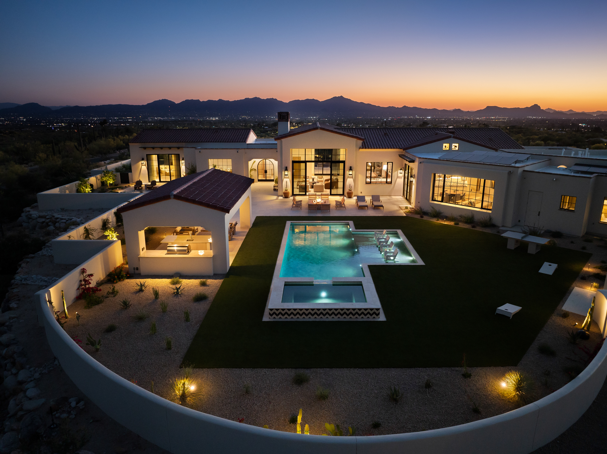 Aerial view at blue hour of a Contemporary Spanish Colonial compound in Dove Mountain, Marana, Arizona — white plaster walls, terracotta clay tile rooflines, illuminated pool, and the valley floor spread out below with mountain silhouettes on the horizon.