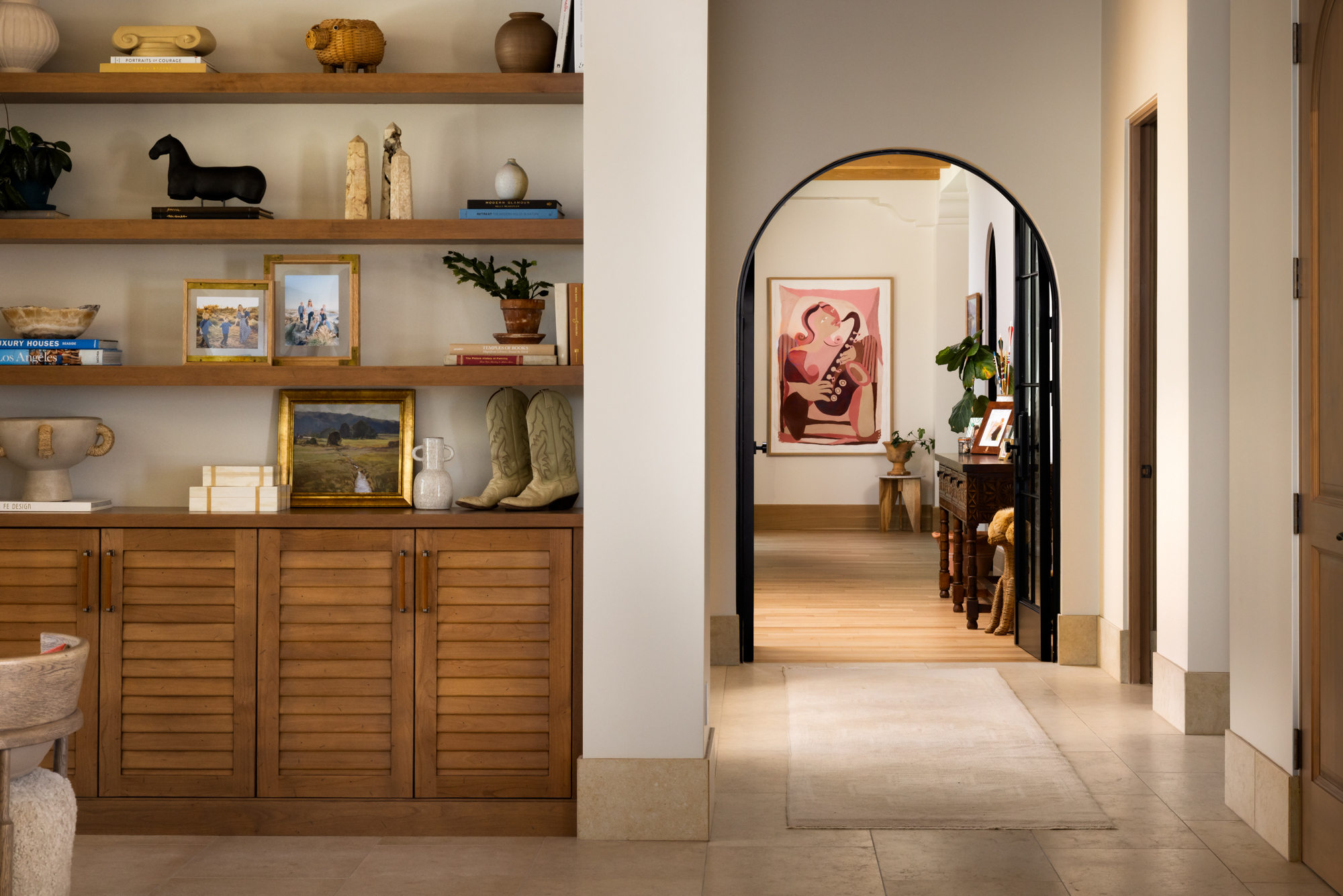 A rounded black-framed arch opens from a shelf-lined alcove into a long corridor at the Dove Mountain home — shelves hold books, pottery, a painting in a gilded frame, and cowboy boots. Art is visible at the far end of the hallway.