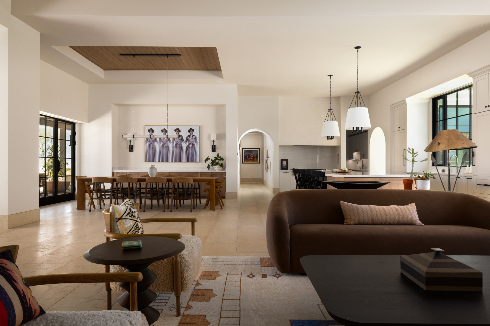 Wide interior view of the great room and dining area at the Dove Mountain home — two arches visible in the line of sight organize the open floor plan, with the kitchen beyond and steel-frame windows at left showing the desert outside.