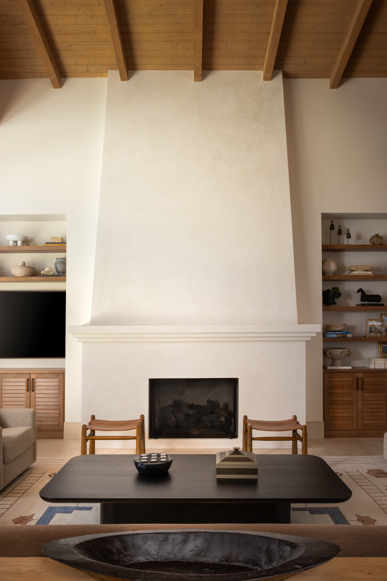 The great room fireplace at the Dove Mountain home — a monumental plaster form running from floor to nearly the full ceiling height, flanked by built-in shelving on both sides, with two leather chairs at the base and exposed timber beams above.
