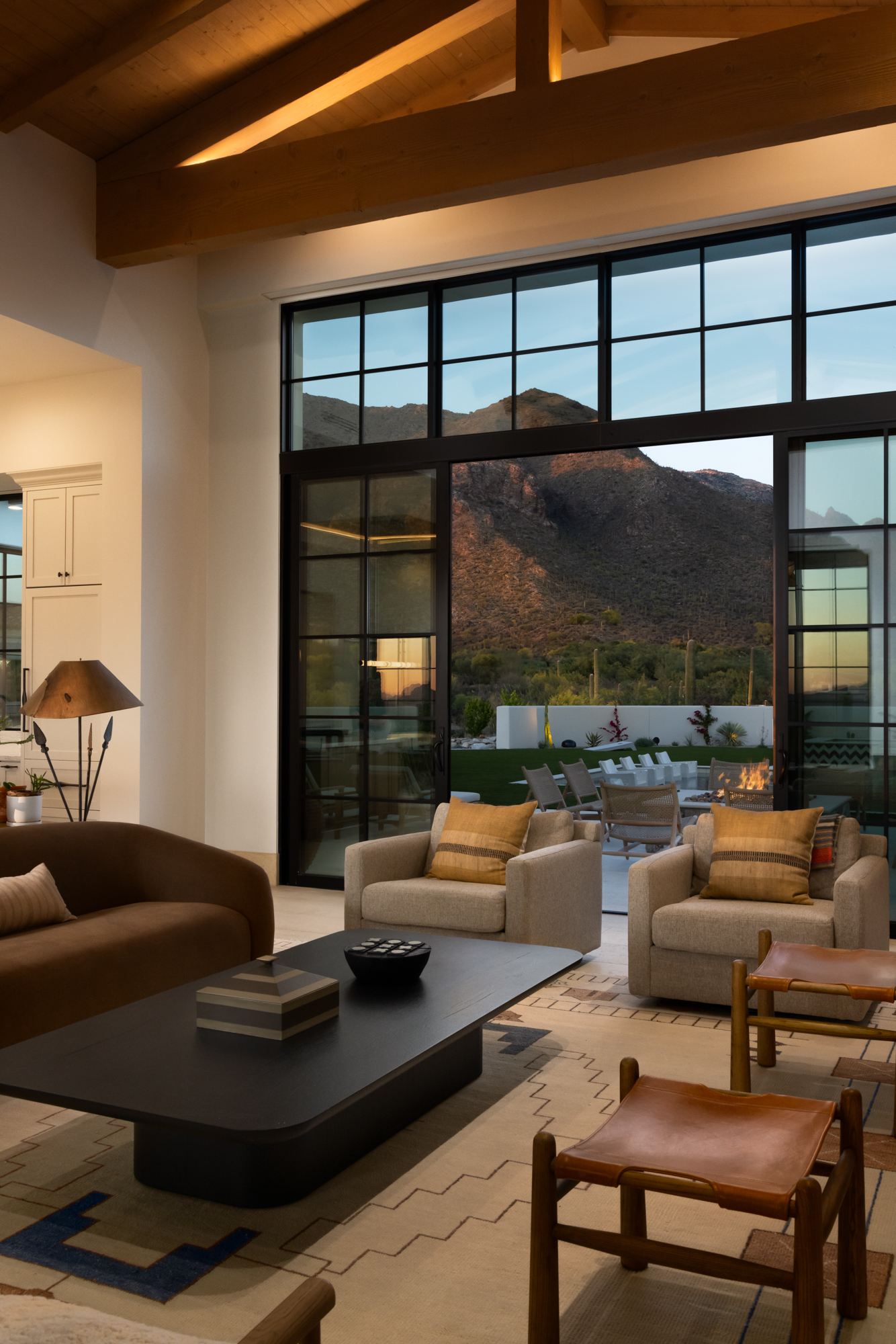 Interior of the Dove Mountain home at dusk — the living room's steel-frame grid windows hold the warm interior against the cooling mountain light outside, with the fire pit glowing in the courtyard beyond and the mountain range at blue hour.