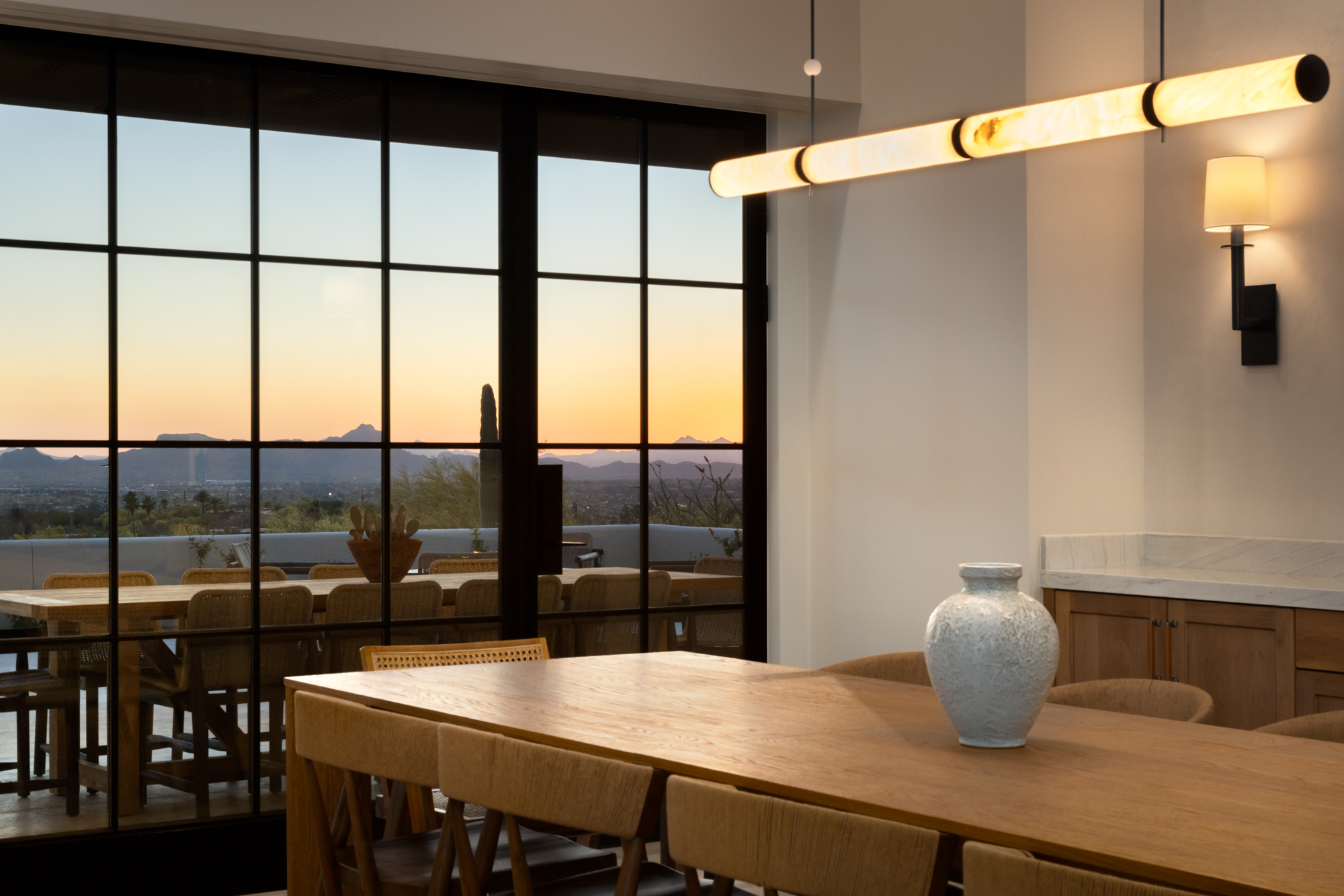 The dining room at the Dove Mountain home at dusk — an alabaster tube pendant glows warm above a wood table, while through the steel-frame grid windows the Tucson valley floor spreads out below with the last amber light on the horizon and mountain silhouettes against the deepening sky.