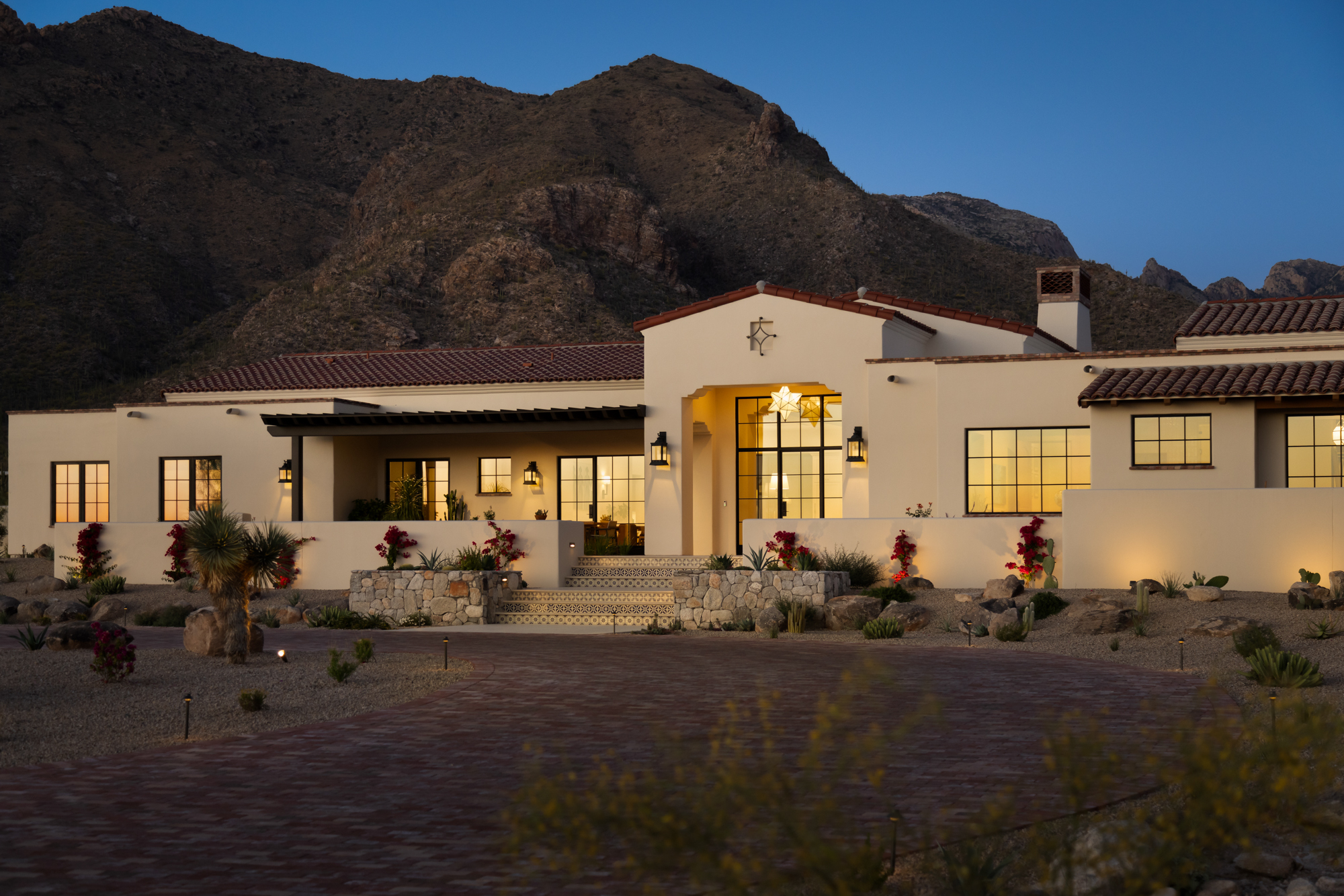 The facade of the Dove Mountain home at blue hour — white plaster, terracotta tile roofline, steel-frame grid windows glowing warm from within, arched entry with lantern fixtures, and the mountain range rising behind in the deep blue dusk sky.