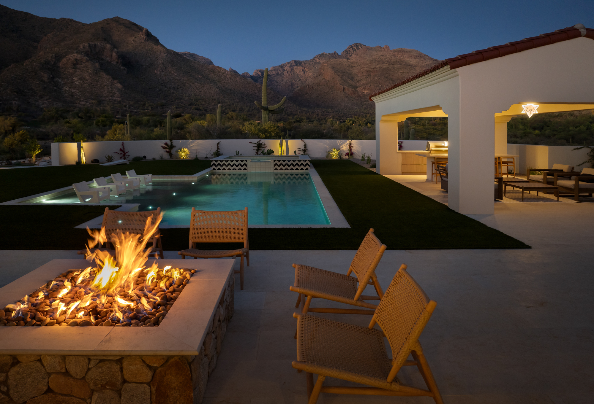 Fire pit in the foreground with active flames, the illuminated pool glowing blue-green beyond, the covered outdoor kitchen to the right lit by lanterns, a saguaro cactus in the middle ground, and the mountain range above in the deep blue night sky.