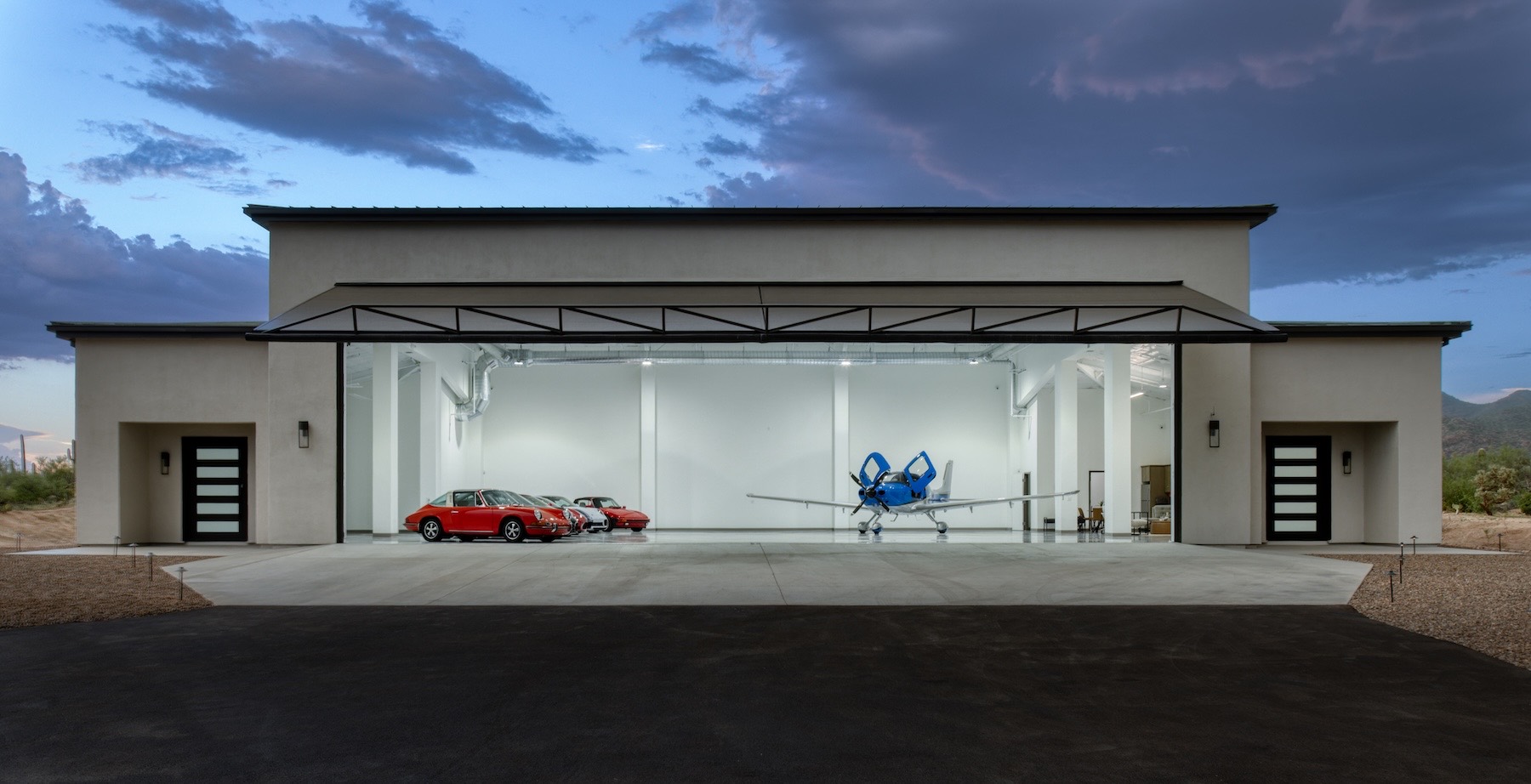 Front elevation of the hangar at twilight — the wide main door raised, the white interior glowing, a red Porsche and two more sports cars at left and a blue-and-white Cirrus at right, under a dramatic blue-purple evening sky.