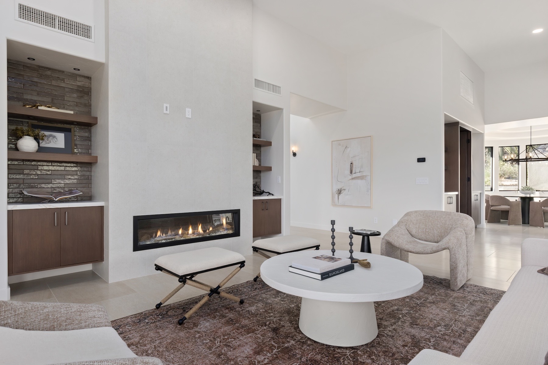 The living room of the home, staged by Elevated Interiors — a linear gas fireplace in a white wall flanked by tile-backed niches with wood shelves, a round white coffee table, a sculptural bouclé chair, and a patterned rug.