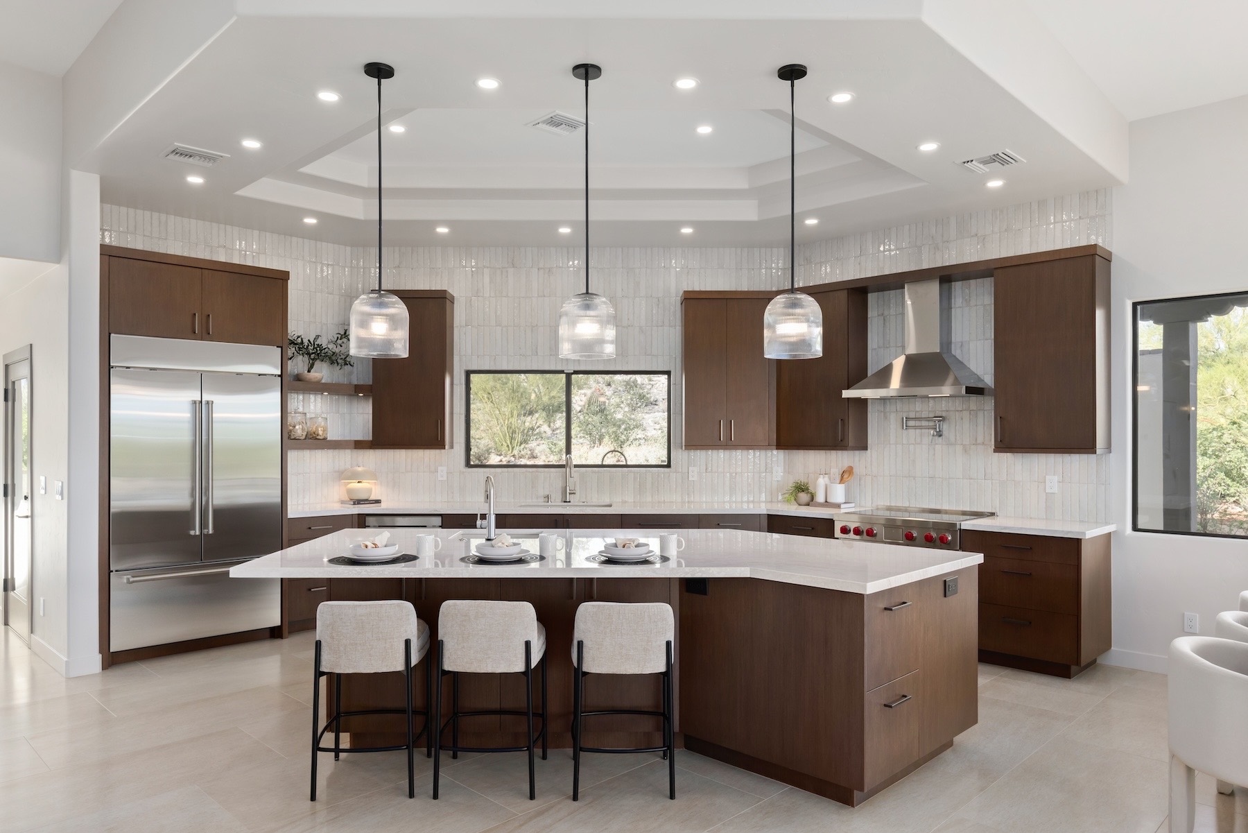 The kitchen of the home — walnut-toned cabinetry, a large white-topped island with three upholstered stools, three glass pendant lights, a stainless range hood, and a tray ceiling with recessed lights.
