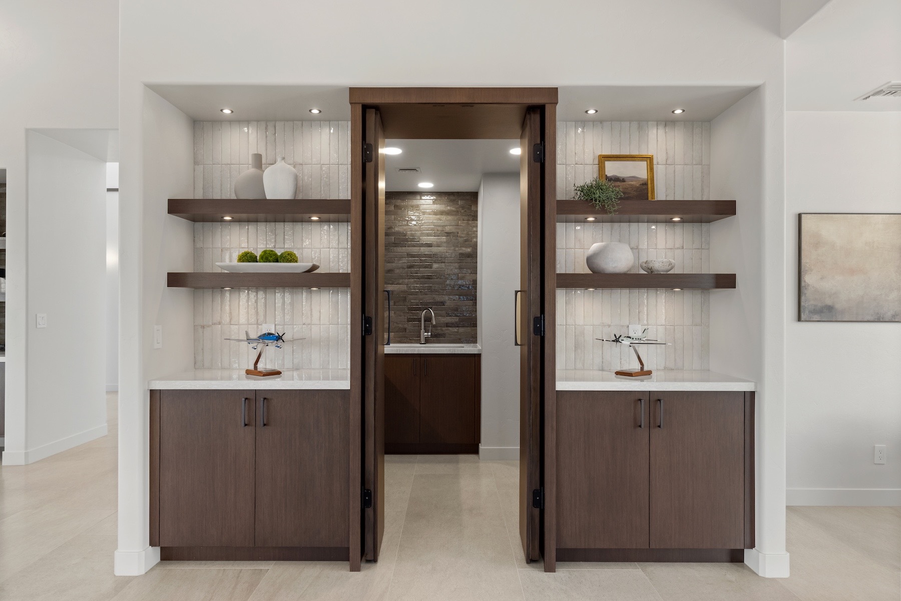 Tall wood double doors open onto a tiled pantry with a sink, set between two lit niches with floating wood shelves holding white ceramics, greenery, a small framed painting, and two model airplanes on stands.