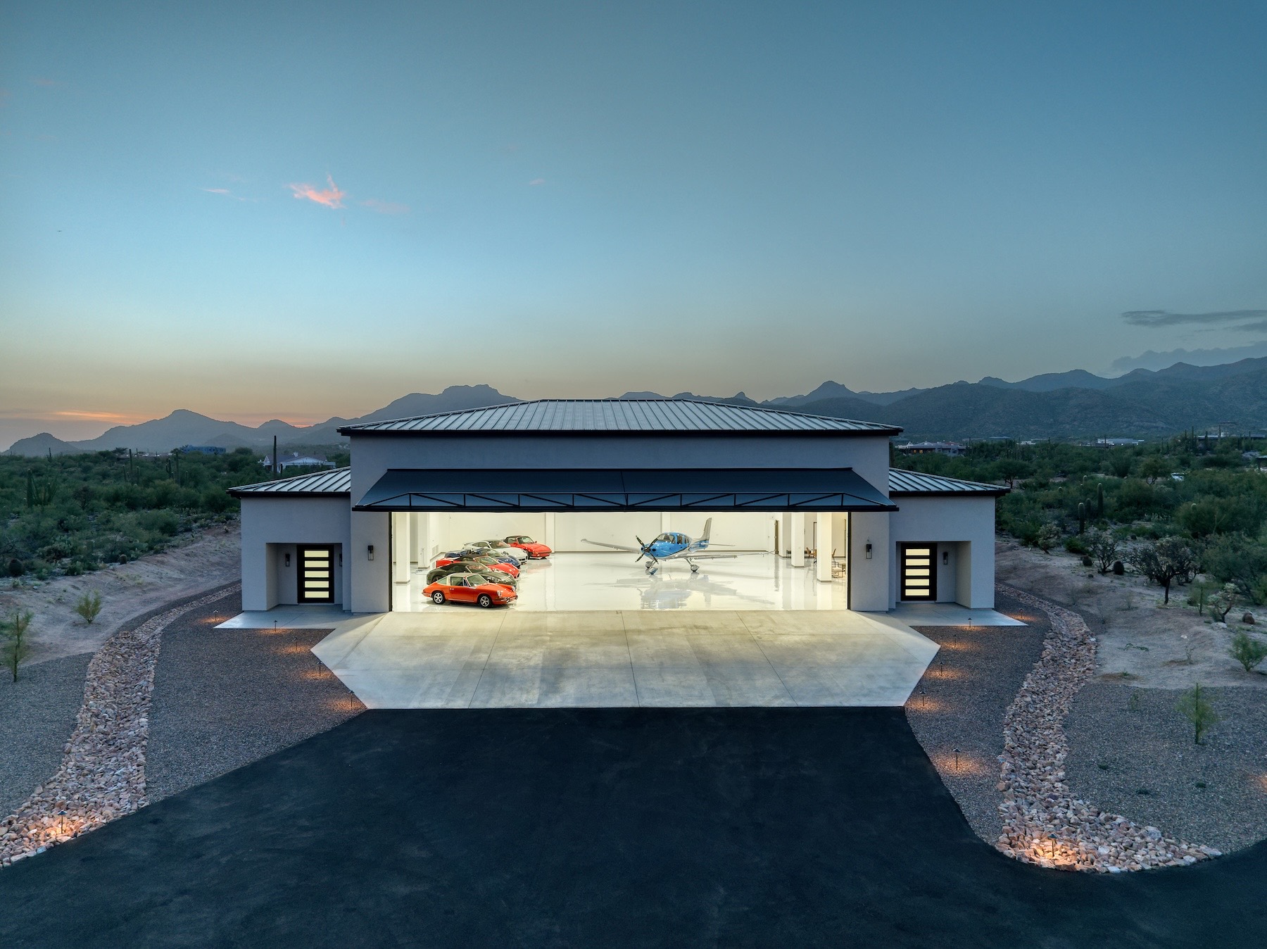 Aerial drone view at twilight of the hangar at La Cholla Airpark — the white building with its main door open and lit from within, a Cirrus and a row of sports cars inside, a wide concrete apron and lit landscape borders in front, and desert and mountains behind under a blue evening sky.