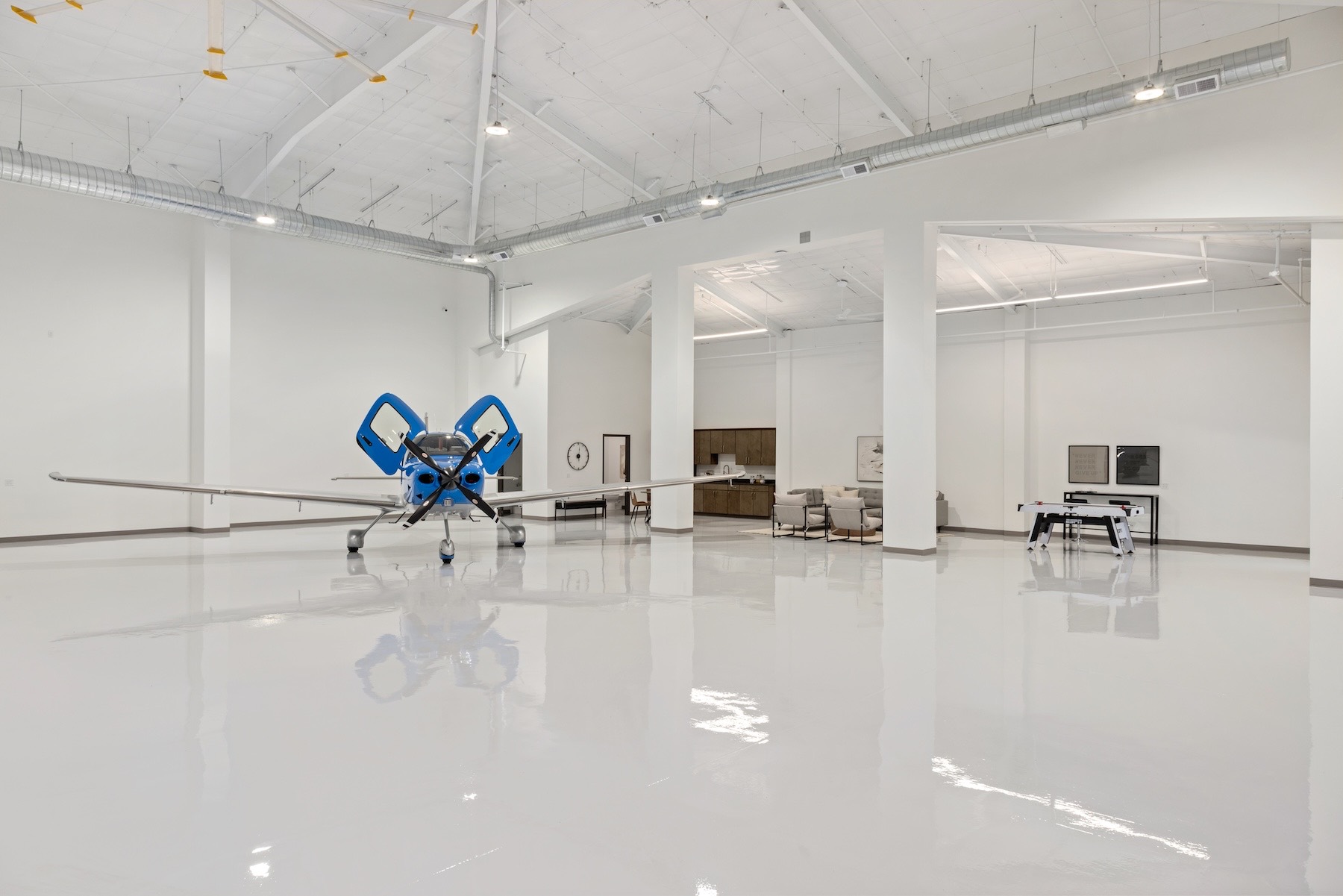 Interior of the 7,000-square-foot hangar at La Cholla Airpark — bright white walls, exposed white trusses, and a glossy white epoxy floor reflecting a blue-and-white Cirrus parked at left, with a staged lounge and kitchenette in the background.