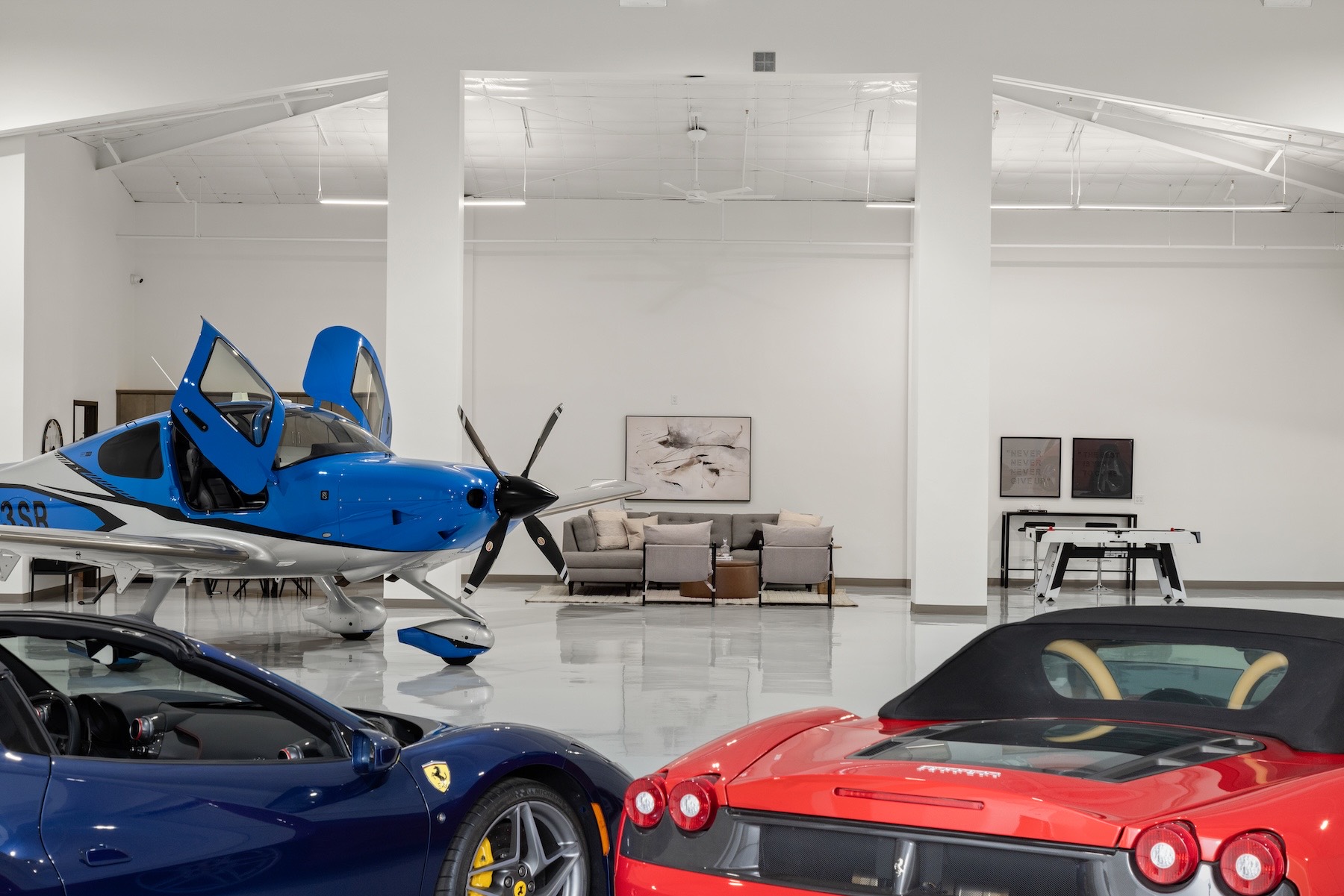 Inside the hangar, a blue Ferrari and a red Ferrari convertible in the foreground frame a blue-and-white Cirrus with its canopy doors open, with the staged hangar lounge and framed artwork beyond.