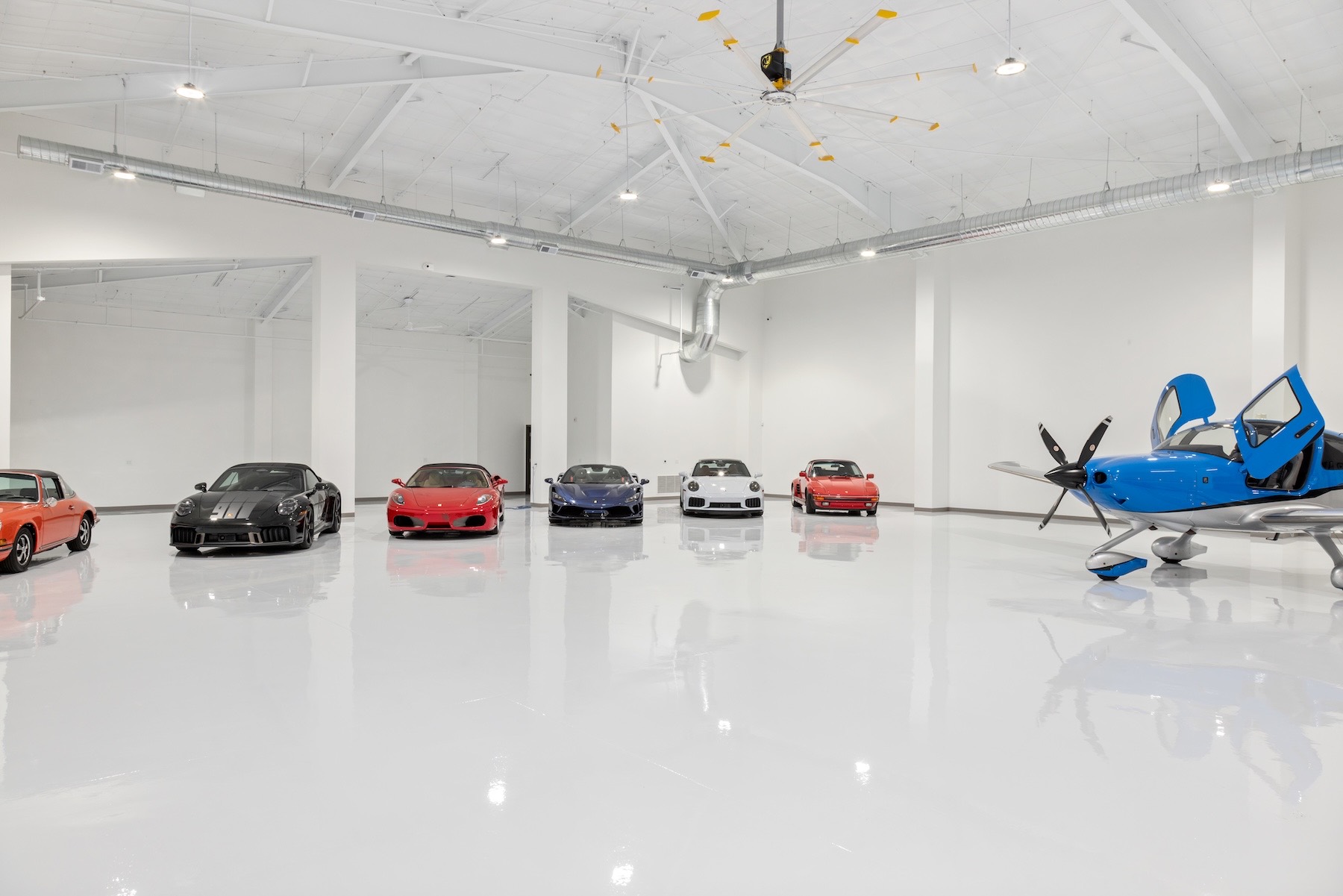 Wide interior of the hangar — six sports cars including Porsches and Ferraris lined up across the glossy white epoxy floor, with the nose of a blue-and-white Cirrus at right, a large ceiling fan and white trusses overhead.