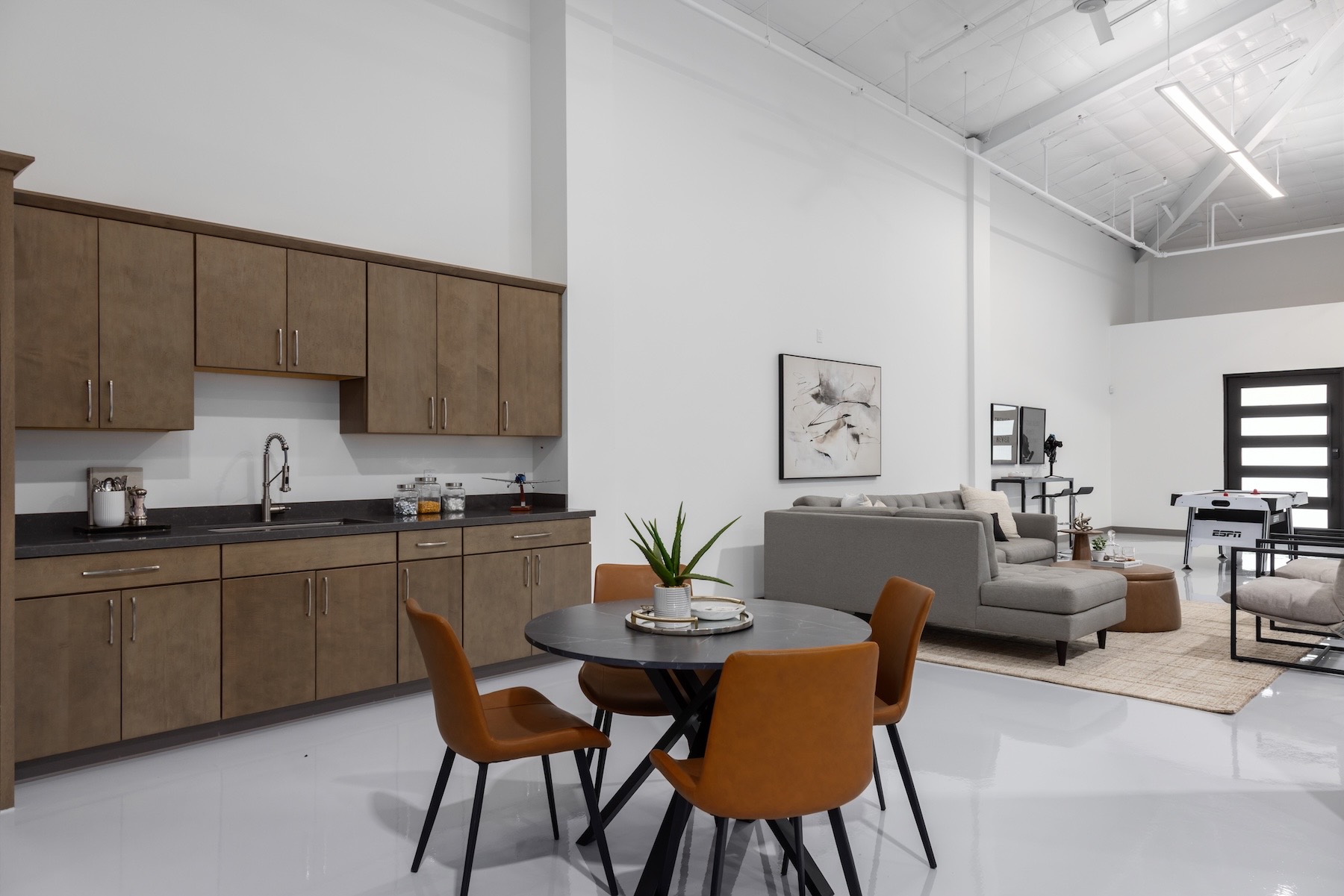 The staged lounge inside the hangar by Elevated Interiors — a kitchenette with wood cabinets, a round dining table with cognac leather chairs, a gray sectional on a woven rug, framed abstract art, and a game table near a modern glass entry door.