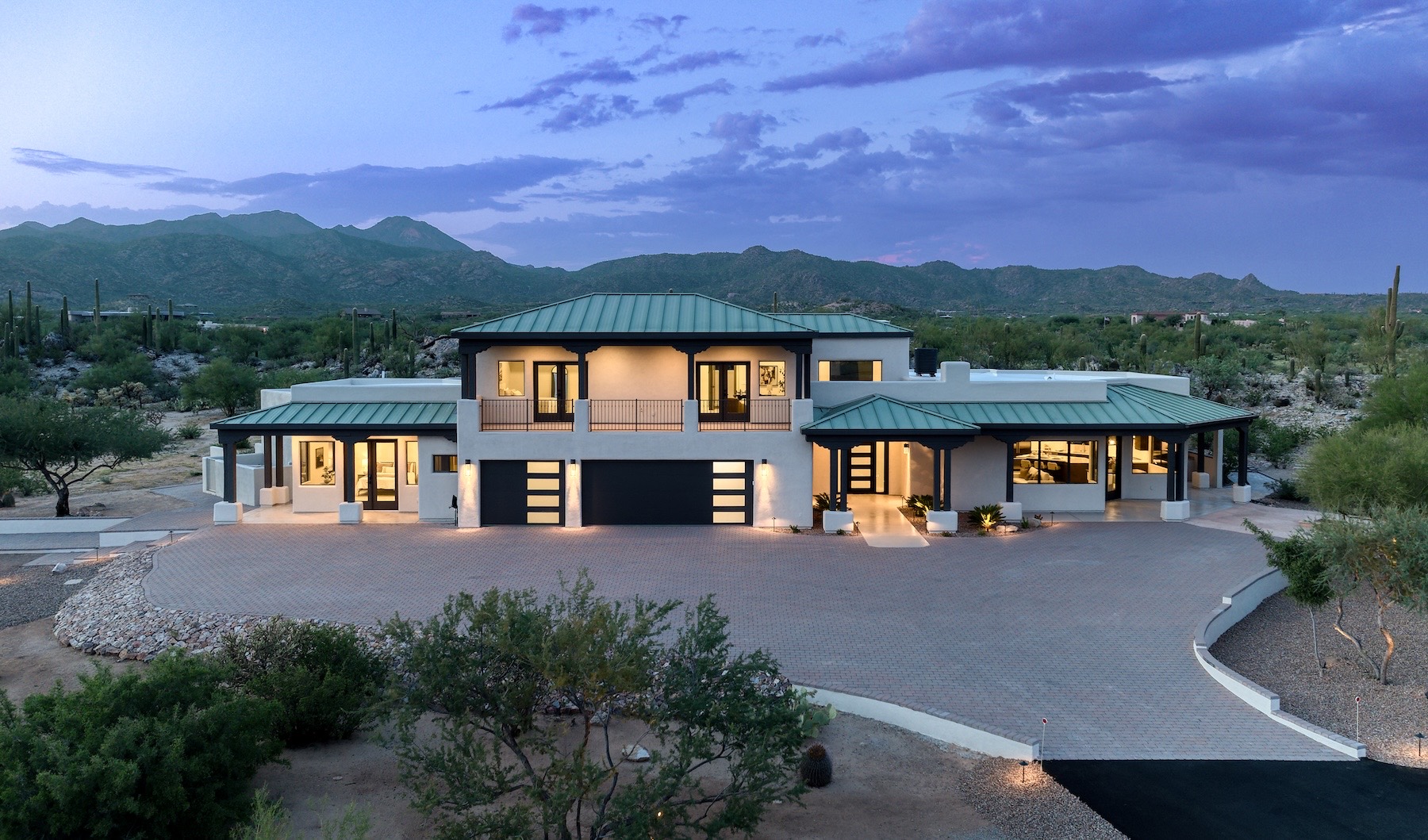 Elevated view at twilight of the home at La Cholla Airpark — a white two-story house with green standing-seam metal roofs, warm light in every window, a wide paver motor court, and desert and mountains beyond under a purple sky.